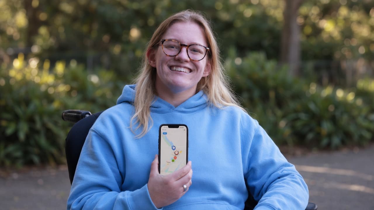 Daisy Hoe smiling outdoors in her wheelchair, holding a phone that shows a colour-coded map